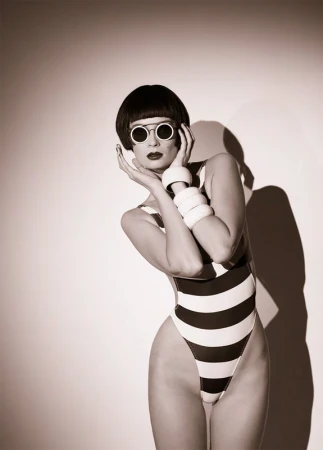 Model with blunt bob haircut wearing round sunglasses and striped swimsuit, posing with hands near face against a plain background