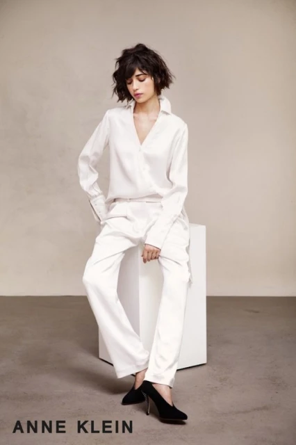 A brunette woman with bangs in a white blouse and trousers poses in a fashion shot. She sits on a white block with black heels.