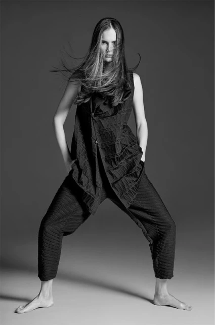 Fashion portrait of a female model with long brown hair, wearing a textured black outfit with loose pants, posing in a studio.