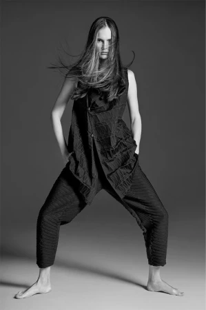 Fashion portrait of a female model with long brown hair, wearing a textured black outfit with loose pants, posing in a studio.