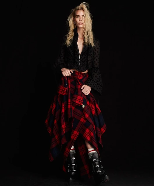 A female fashion model poses in a black top and red plaid skirt with ski boots in a studio setting against a black backdrop.