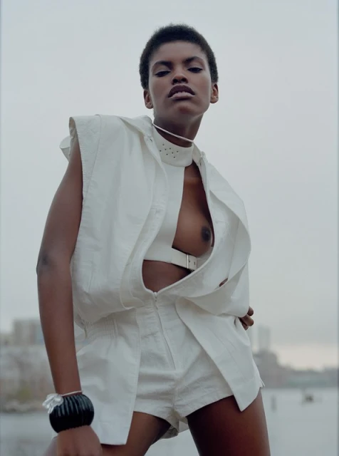 Adult female model with short dark hair poses in a white shorts outfit with a river and buildings in the distant background.