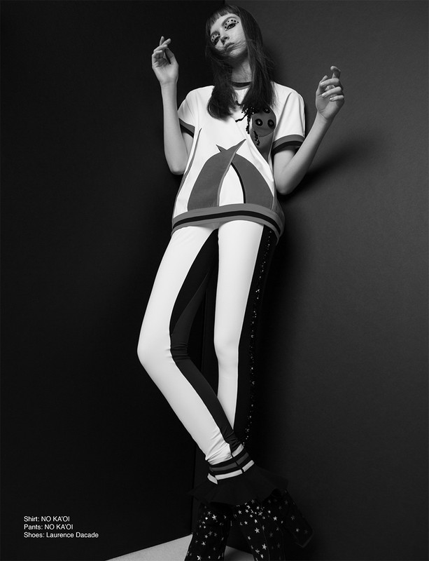 Black-and-white studio shot of a stylish young woman with bold eye makeup, wearing a graphic t-shirt, striped pants, and star-patterned boots.