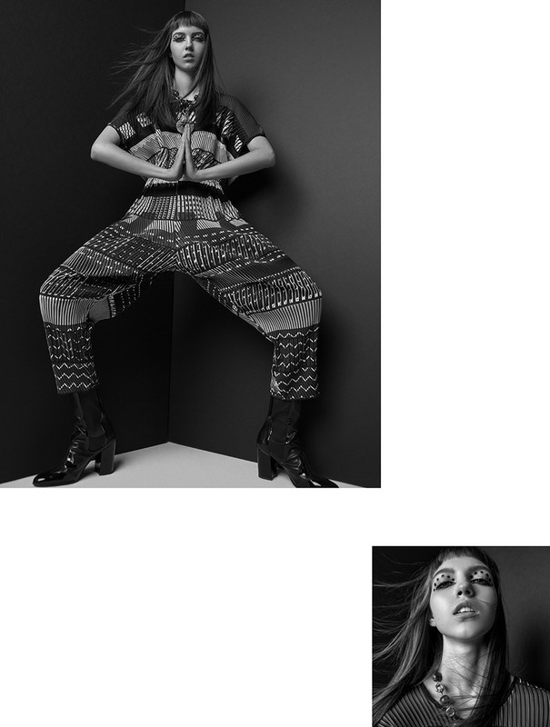 A young adult female model poses in a black and white studio shot wearing a patterned top and pants with black boots, expressive makeup.