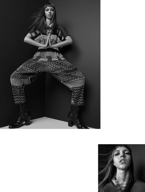 A young adult female model poses in a black and white studio shot wearing a patterned top and pants with black boots, expressive makeup.