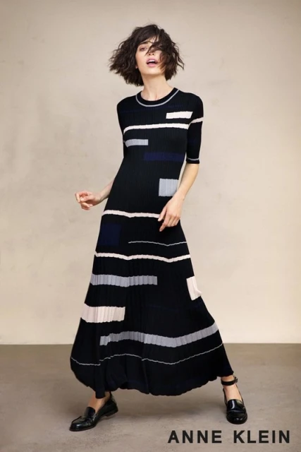 A slender woman in a patterned black dress stands in a neutral studio, her gaze off-center, showing off a contemporary and elegant look.
