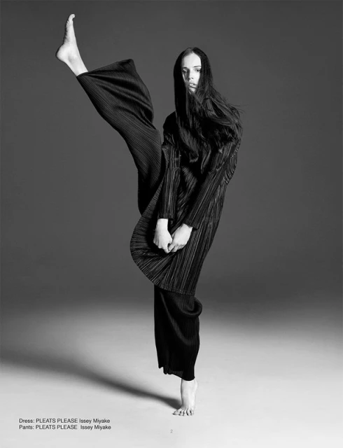 Monochrome fashion shot of a young woman in pleated black top and trousers, striking a dynamic pose with one leg raised in a studio setting.