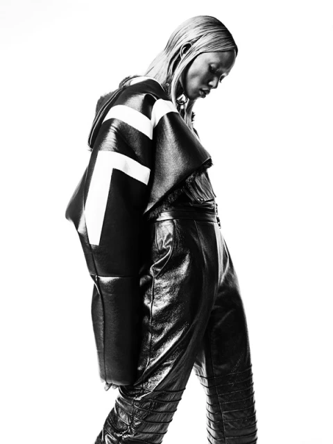 Editorial fashion shot of a Black female model wearing a black and white jacket and shiny black leather pants against a white background.
