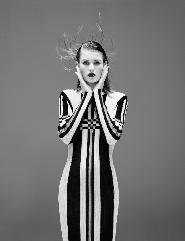 Portrait of a woman in a black and white patterned dress with hands near her face against a grey background in a studio setting.