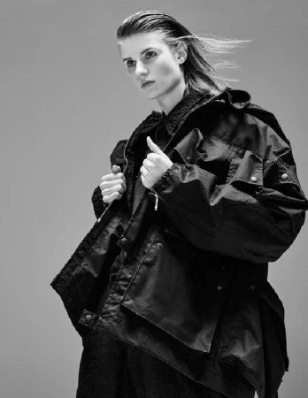 Model with slicked-back hair wearing a dark utility jacket against a plain grey studio backdrop