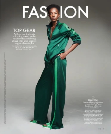 Model standing in a green satin track suit against a grey studio backdrop featuring large white editorial magazine typography