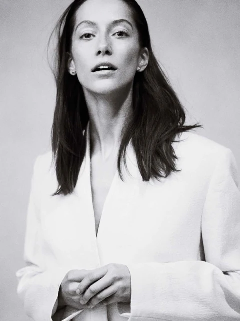Monochrome portrait of an adult female with long hair, wearing a light-colored blazer, standing in front of a neutral background.