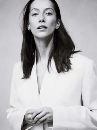 Monochrome portrait of an adult female with long hair, wearing a light-colored blazer, standing in front of a neutral background.