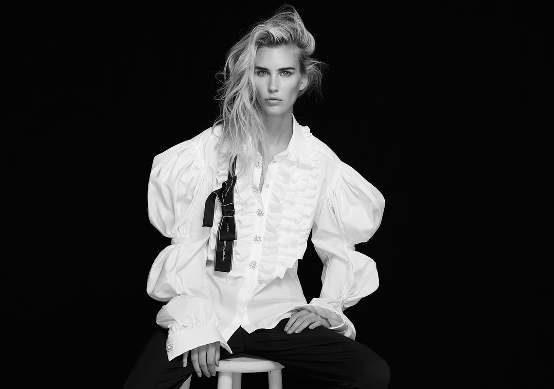 Blonde woman in a white ruffled shirt and dark pants poses against a black backdrop, seated on a stool in a fashion portrait.