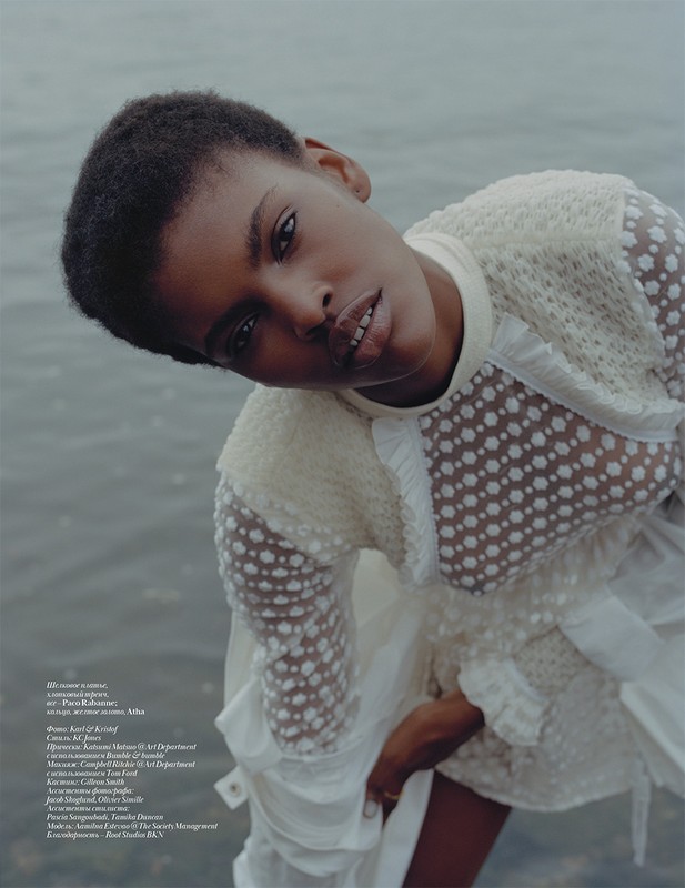 Fashion editorial shot of a Black female model in a white lace dress posing outdoors near water with a soft, muted colour palette.