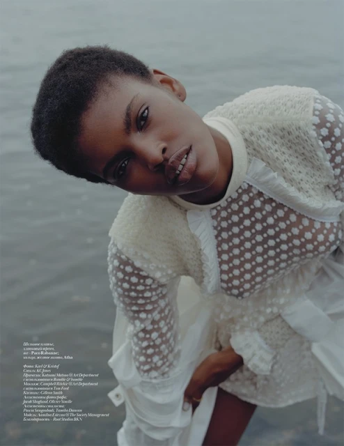 Fashion editorial shot of a Black female model in a white lace dress posing outdoors near water with a soft, muted colour palette.