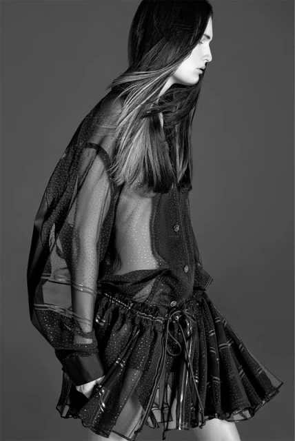 Black-and-white fashion shot of a slender person with long hair wearing a sheer black dotted top and skirt against a grey background.