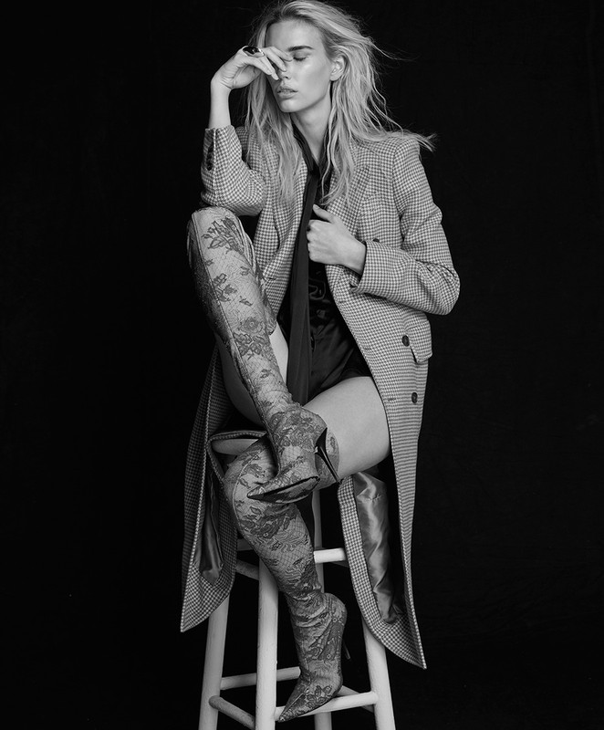 Monochrome studio portrait of a woman in thigh-high lace boots, a coat, and a dark shirt, sitting on a white stool against a black background.