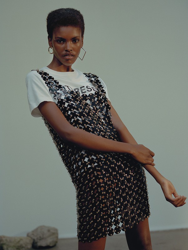 An adult woman poses in a sleeveless, open-weave black dress with a white t-shirt against a neutral studio backdrop.