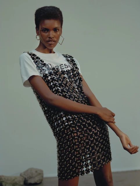 An adult woman poses in a sleeveless, open-weave black dress with a white t-shirt against a neutral studio backdrop.