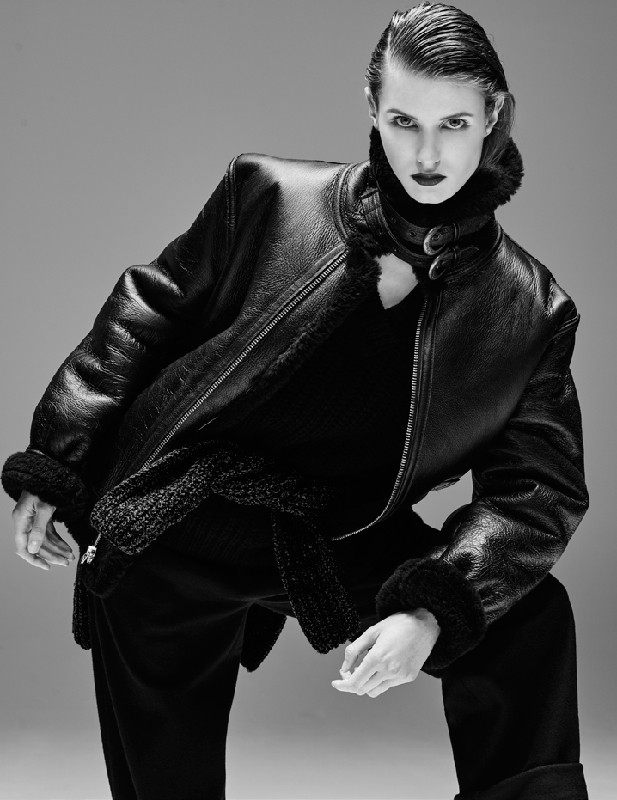 Monochrome portrait of an adult female model in a black shearling bomber jacket, black top and pants in a studio setting with gray backdrop.