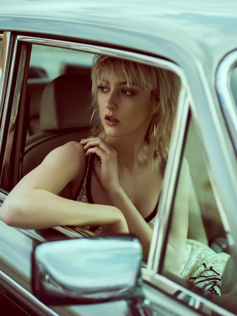 A blonde woman inside a vintage car with a cream-colored interior, hoop earrings, and spaghetti strap top looks toward the window.