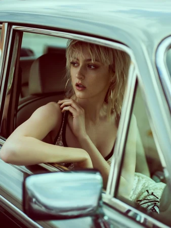 A blonde woman inside a vintage car with a cream-colored interior, hoop earrings, and spaghetti strap top looks toward the window.
