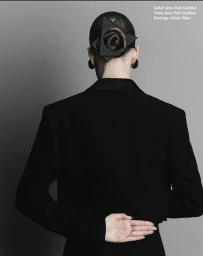 Rear view of a person with dark hair in a knotted bun wearing a black blazer and dark earrings against a grey background.