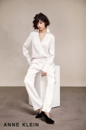 A woman in a white silk blouse and trousers seated on a white block against a grey studio backdrop, wearing black shoes.
