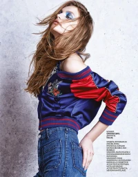 A female model wearing a blue satin bomber jacket with red sleeves and denim jeans against a textured grey concrete wall.