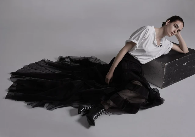 Model wearing a white t-shirt and black tulle skirt leaning against a dark wooden box on a grey seamless background