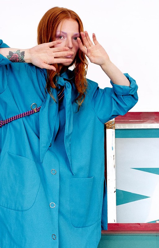 Red-haired woman in a turquoise jacket poses with hands near her face in front of a white backdrop, next to a painted surface.