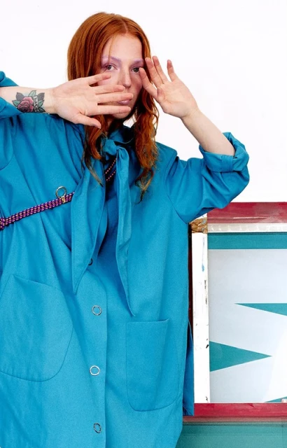 Red-haired woman in a turquoise jacket poses with hands near her face in front of a white backdrop, next to a painted surface.