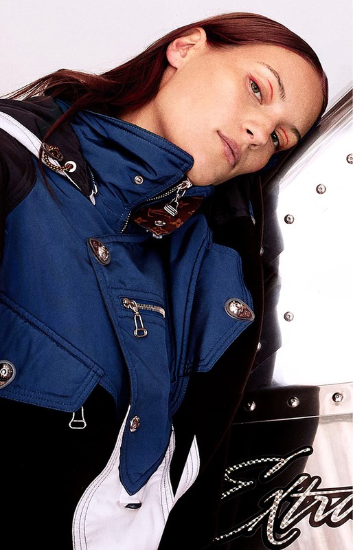 Model with red hair leaning against a metallic trunk wearing a blue, black, and white hooded jacket styled by Keiko Hamaguchi.