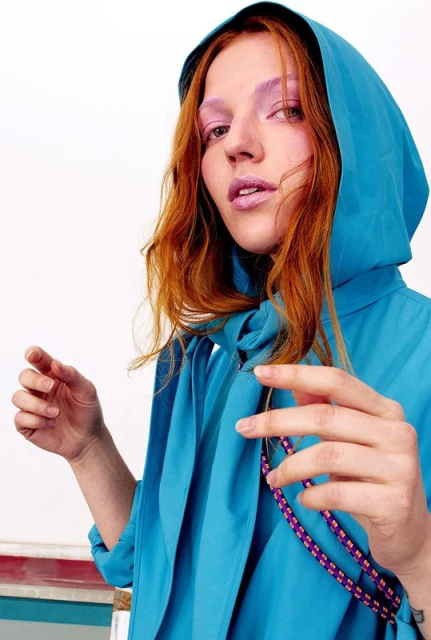 A red-haired model in a hooded turquoise jacket gazes directly at the camera in a studio shot, with bright makeup and a rope necklace.