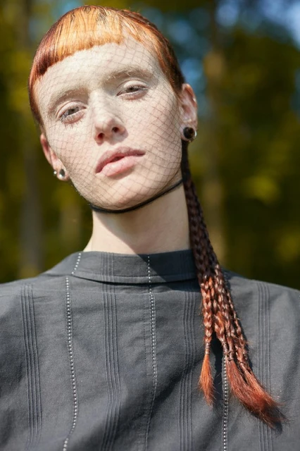 Close-up portrait of a woman with reddish hair, a net veil over her face, and a collared shirt, outdoors in natural light.
