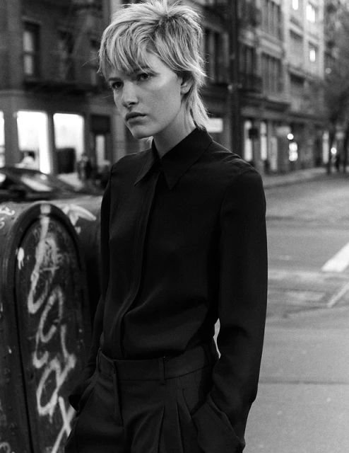 Black and white portrait of a woman with short blonde hair wearing a black blouse and trousers standing on a city street.