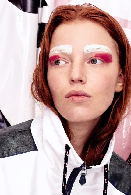 Close-up shot of a redhead adult female fashion model with white and pink painted eyebrows, wearing a sporty white hooded jacket.