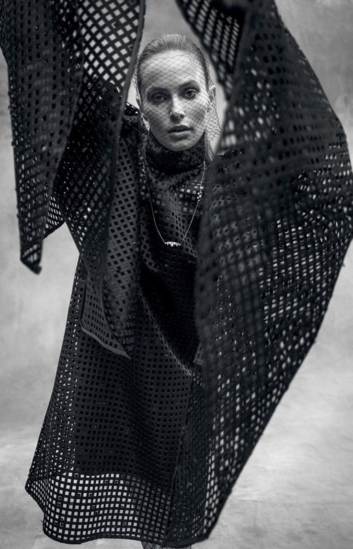 A fashion shot of a woman wearing a black net-like dress and head covering, posing indoors with soft, diffused lighting and a neutral backdrop.