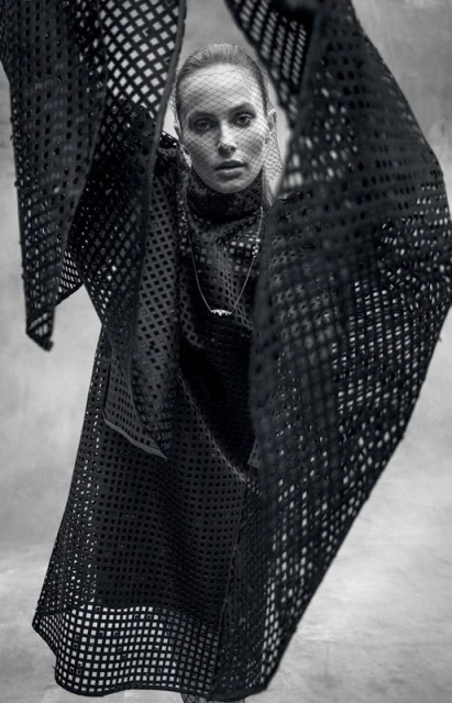 A fashion shot of a woman wearing a black net-like dress and head covering, posing indoors with soft, diffused lighting and a neutral backdrop.