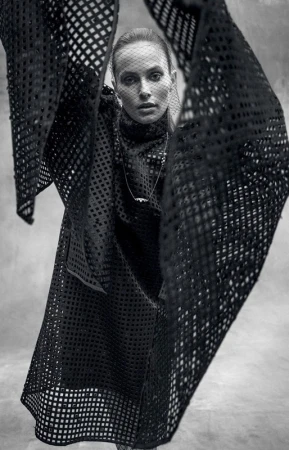 A fashion shot of a woman wearing a black net-like dress and head covering, posing indoors with soft, diffused lighting and a neutral backdrop.