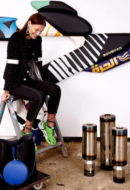 Fashionable woman sits on a ladder in an art studio, wearing a black jacket, stylish pants, green sneakers, and statement earrings.