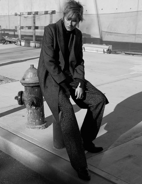 Woman in overcoat sits on fire hydrant outdoors, wearing wide-leg trousers and black top, set against an industrial wall backdrop.