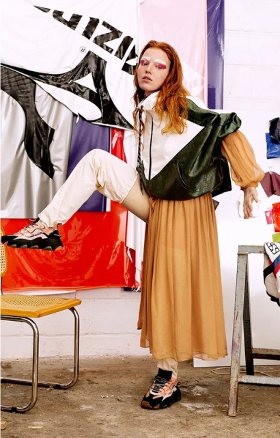 Fashionable woman poses in color-blocked jacket, orange skirt, and platform sneakers near an artist studio with colorful backdrops.