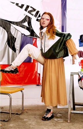 Fashionable woman poses in color-blocked jacket, orange skirt, and platform sneakers near an artist studio with colorful backdrops.