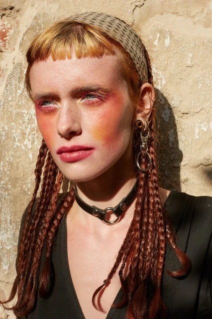 Close-up of a red-haired adult female with an orange and red face makeup, wearing a headscarf and black choker necklace against a beige background.