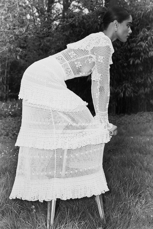 A woman in a flowing, crochet lace dress poses in a grassy outdoor setting, leaning on a stool with foliage in the background.
