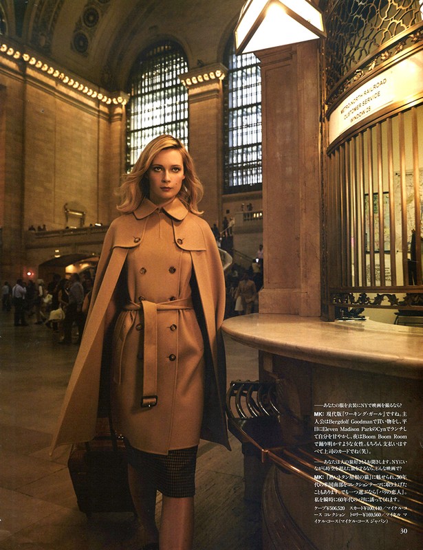Woman in a tan trench coat stands inside Grand Central Terminal, New York City. Large arched windows and a marble ticket booth.