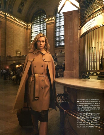 Woman in a tan trench coat stands inside Grand Central Terminal, New York City. Large arched windows and a marble ticket booth.