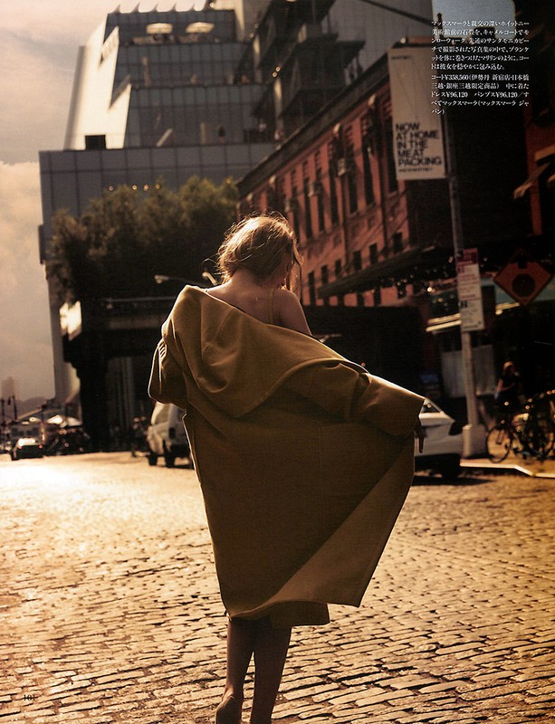 A woman with blonde hair is walking down a cobblestone street in a city, wearing a tan coat, with modern buildings in the background.